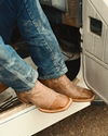 A person wearing blue jeans adjusts their pant leg over brown cowboy boots while standing on dry grass.