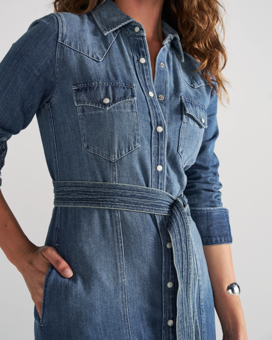 A woman wearing a long-sleeved denim shirt dress with a tie waist, snap buttons, and chest pockets.