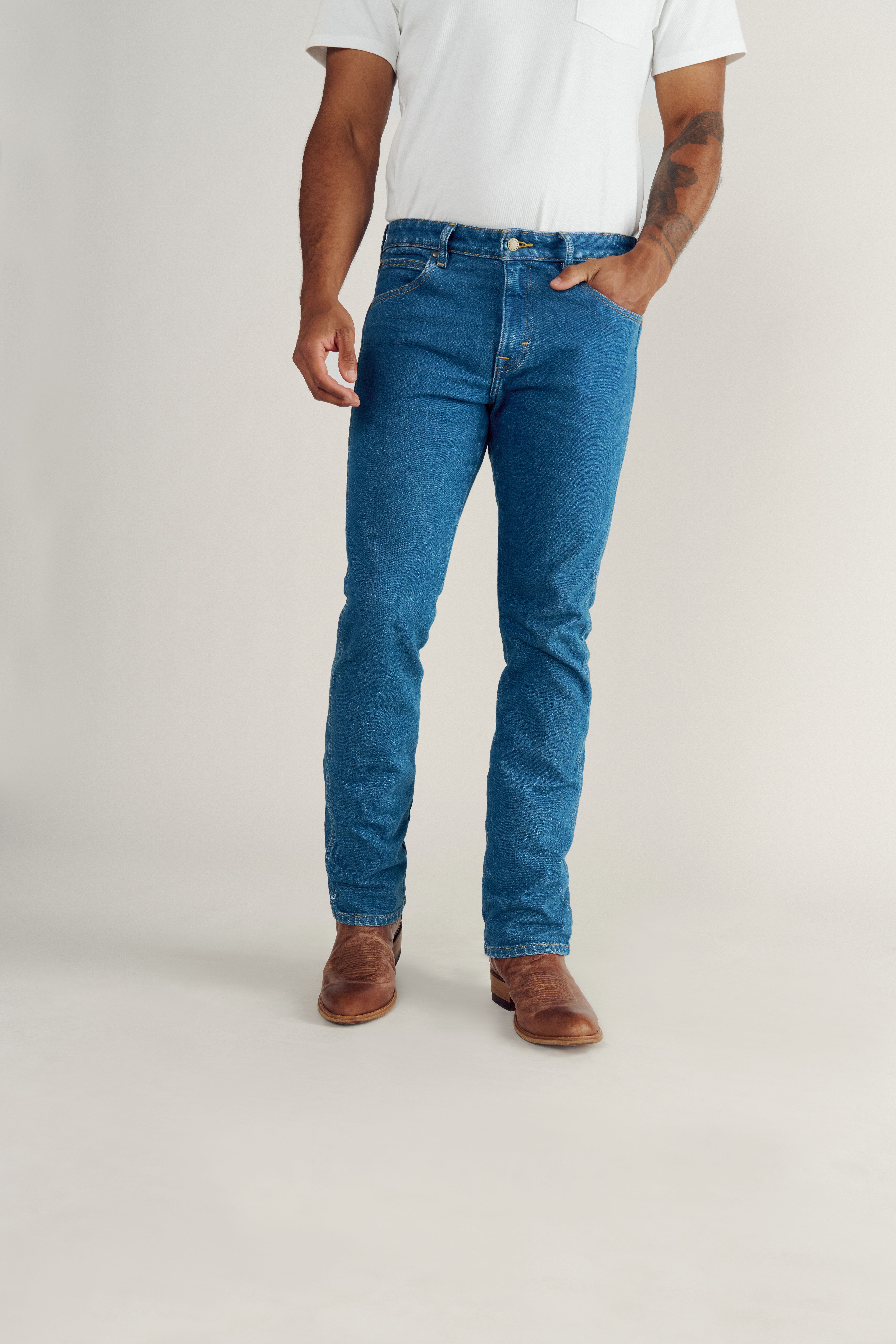 A person wearing a white short-sleeve shirt, blue jeans, and brown boots stands with one hand in their pocket against a plain background.