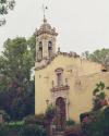 A weathered yellow stone building with a bell tower and arched doorway, surrounded by greenery and flowering plants.