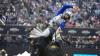 A rodeo rider in safety gear holds onto a bucking bull with one hand raised, surrounded by a blurred audience in an arena.