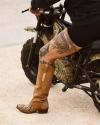 Person with tattooed legs wearing brown cowboy boots sits on a small black motorcycle, with one foot resting on the ground and sand visible beneath.