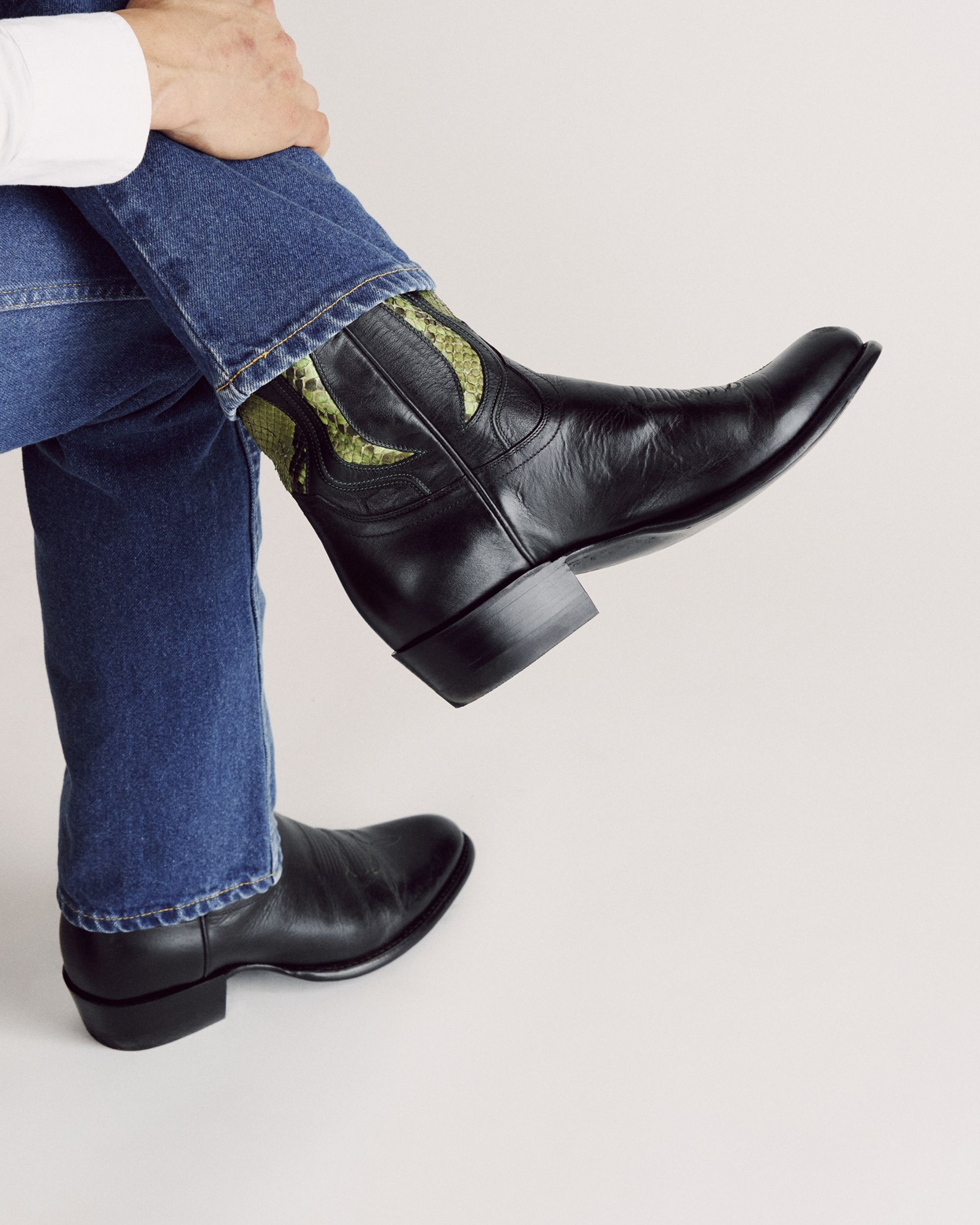 Person wearing blue jeans and black leather cowboy boots with green detailing, sitting with one leg crossed over the other against a plain background.