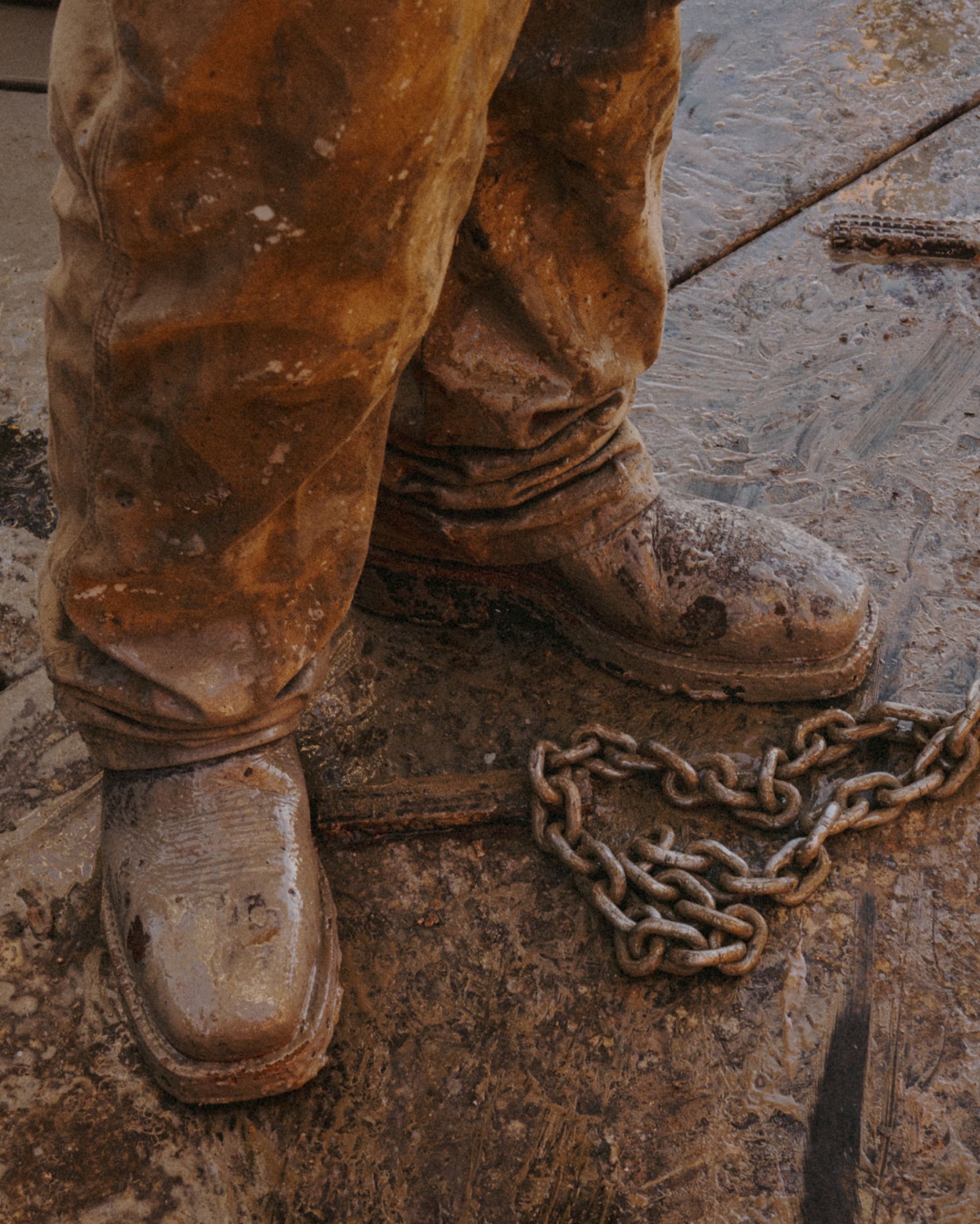 A person wearing muddy work boots and pants stands on a dirty surface next to a metal chain.