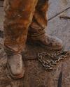 A person wearing muddy work boots and pants stands on a dirty surface next to a metal chain.