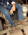 The Rugged Chelsea in Canteen Cowhide. A person in jeans, boots, and a plaid shirt sits on the front bumper of a green Jeep, holding work gloves and a sturdy canteen, with their legs crossed.