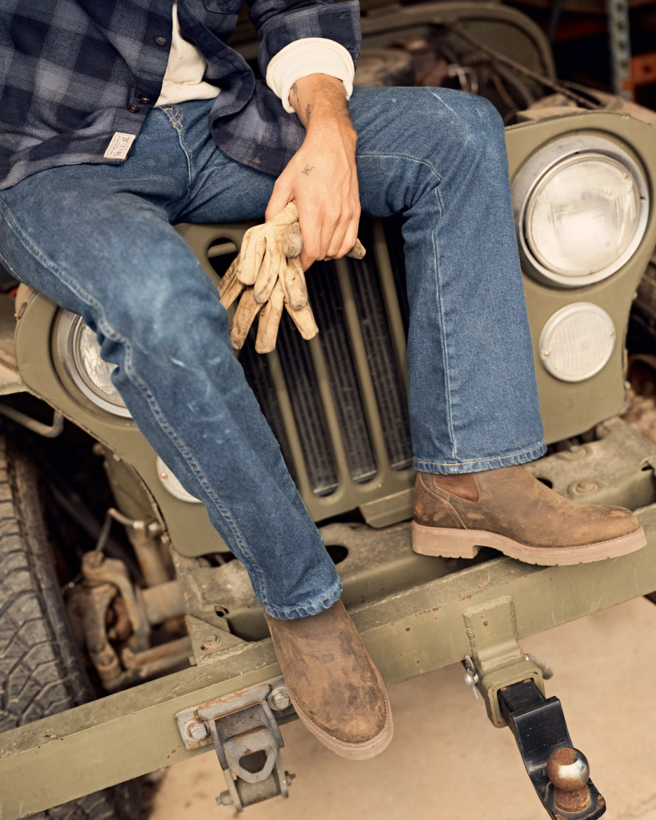The Rugged Chelsea in Canteen Cowhide. A person in jeans, boots, and a plaid shirt sits on the front bumper of a green Jeep, holding work gloves and a sturdy canteen, with their legs crossed.