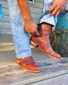 A person wearing blue jeans pulls on a brown leather boot while standing on a wooden porch.