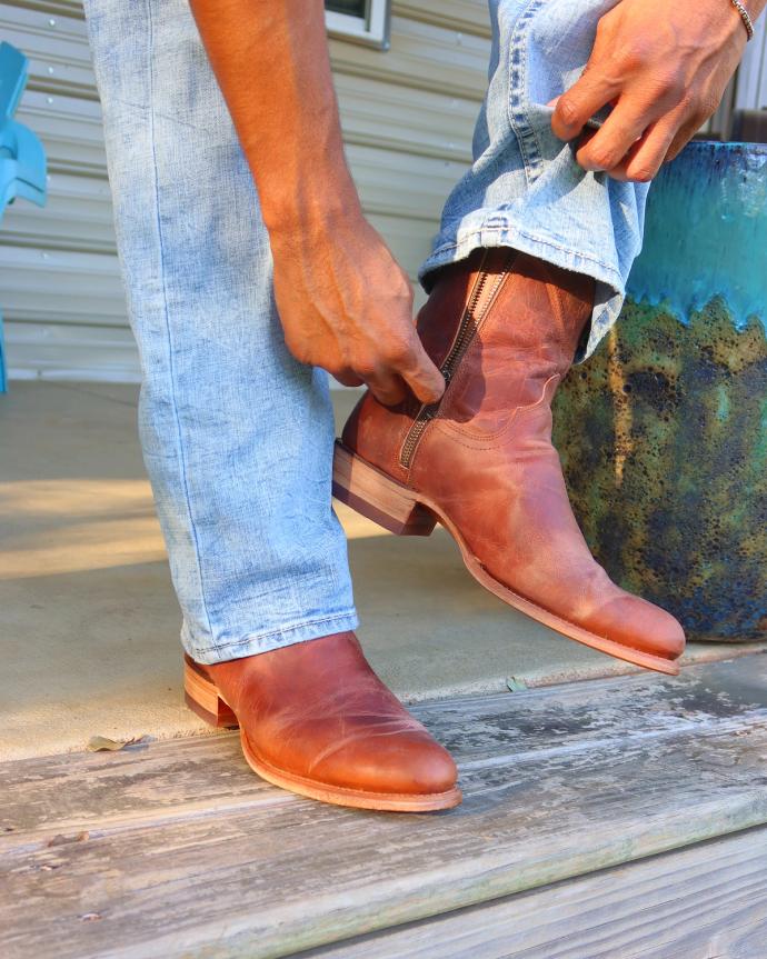 A person wearing blue jeans pulls on a brown leather boot while standing on a wooden porch.