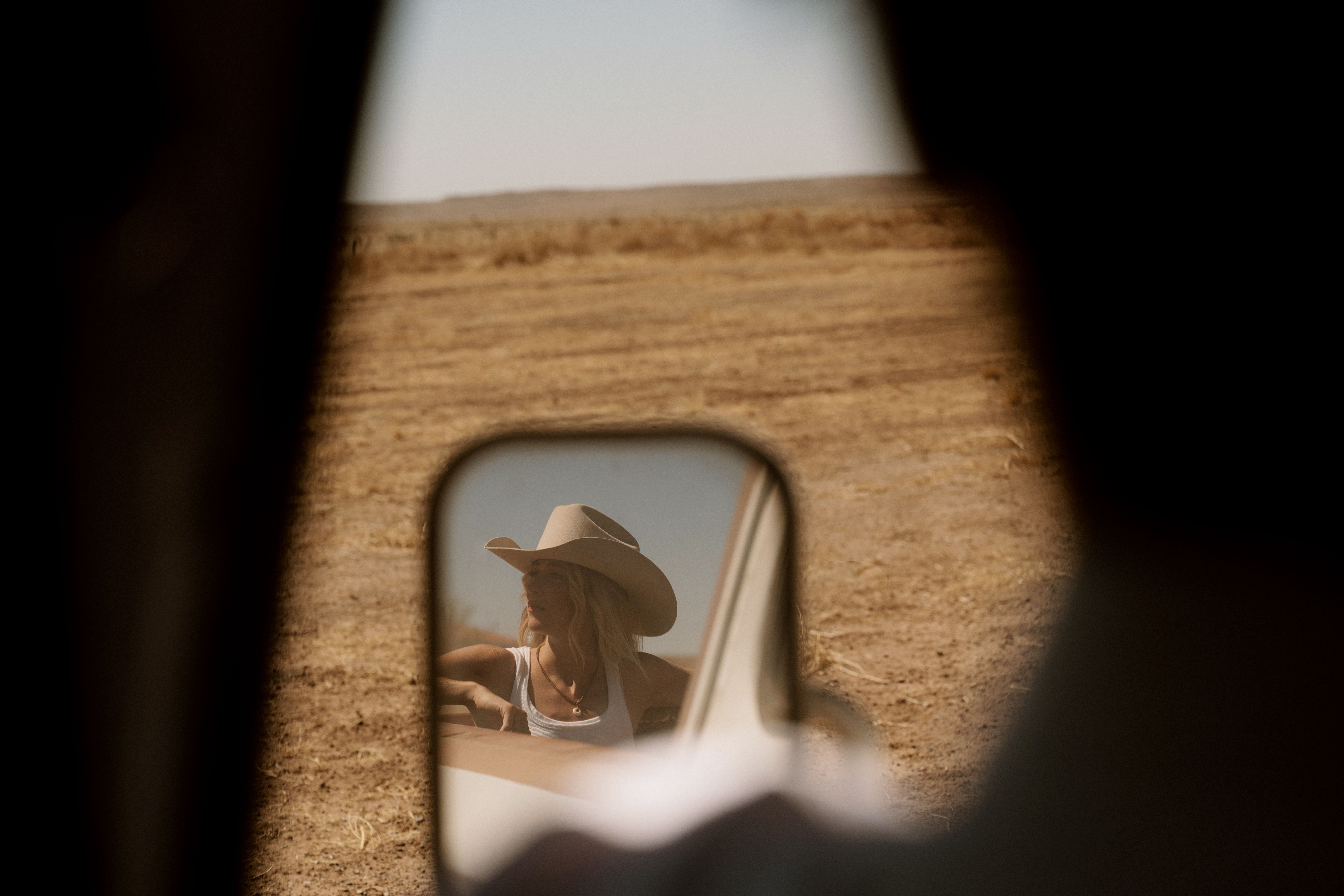 A woman in a cowboy hat stands outside in a dry field, reflected in a vehicle's side mirror, with the vehicle's interior partially visible in the foreground.