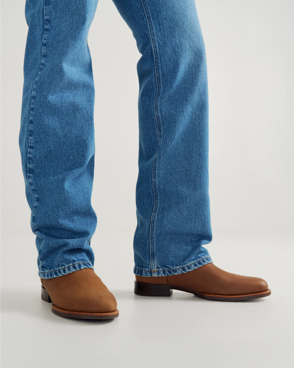 The Glen in Khaki Cowhide. Person wearing blue jeans and brown leather boots, standing on a light-colored surface, showcases classic style with a hint of cowhide charm.