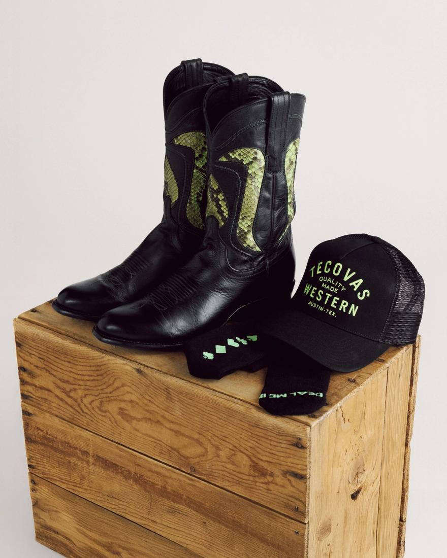 A pair of black cowboy boots with green designs, a black trucker hat, and a pair of black socks arranged on a wooden crate.