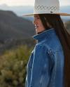 Profile view of a woman with long dark hair wearing a denim jacket and a white hat, standing outdoors with mountains in the background.