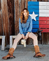 A person in a denim jacket and boots sits in front of a wooden wall painted with the texas flag.