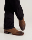 A person wearing dark pants and brown leather dress boots stands with one foot slightly raised against a plain background.