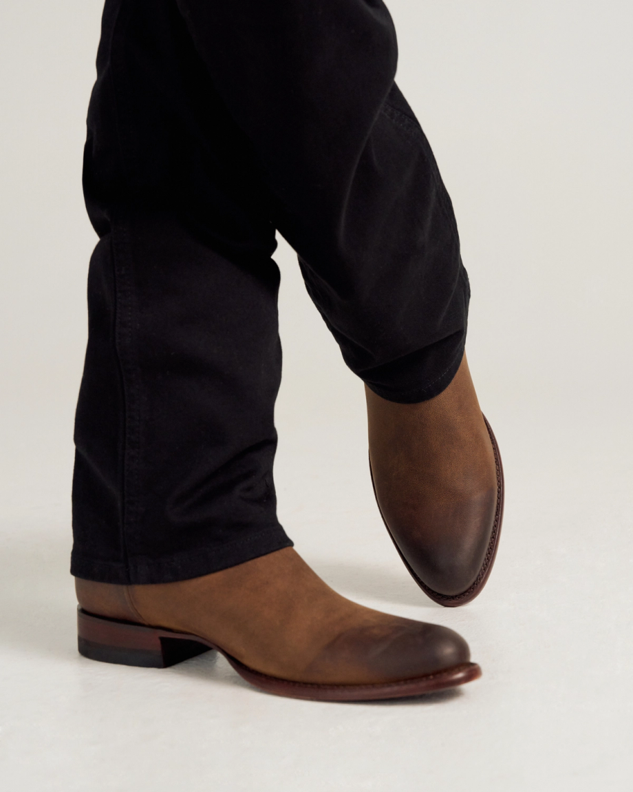 A person wearing dark pants and brown leather dress boots stands with one foot slightly raised against a plain background.