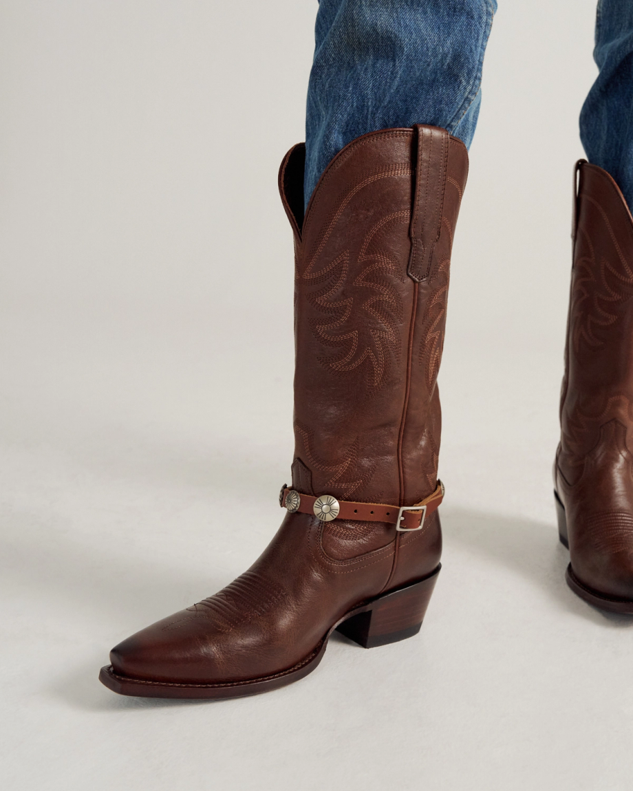 A person wearing blue jeans and a pair of brown leather cowboy boots with a decorative strap around the ankle.