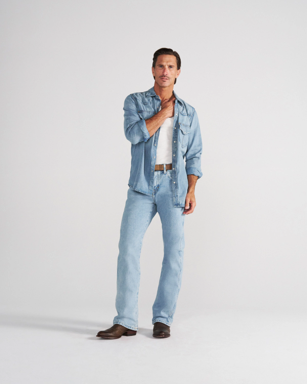 A man wearing a light-wash denim shirt over a white tank top, light-wash blue jeans, and brown leather boots.
