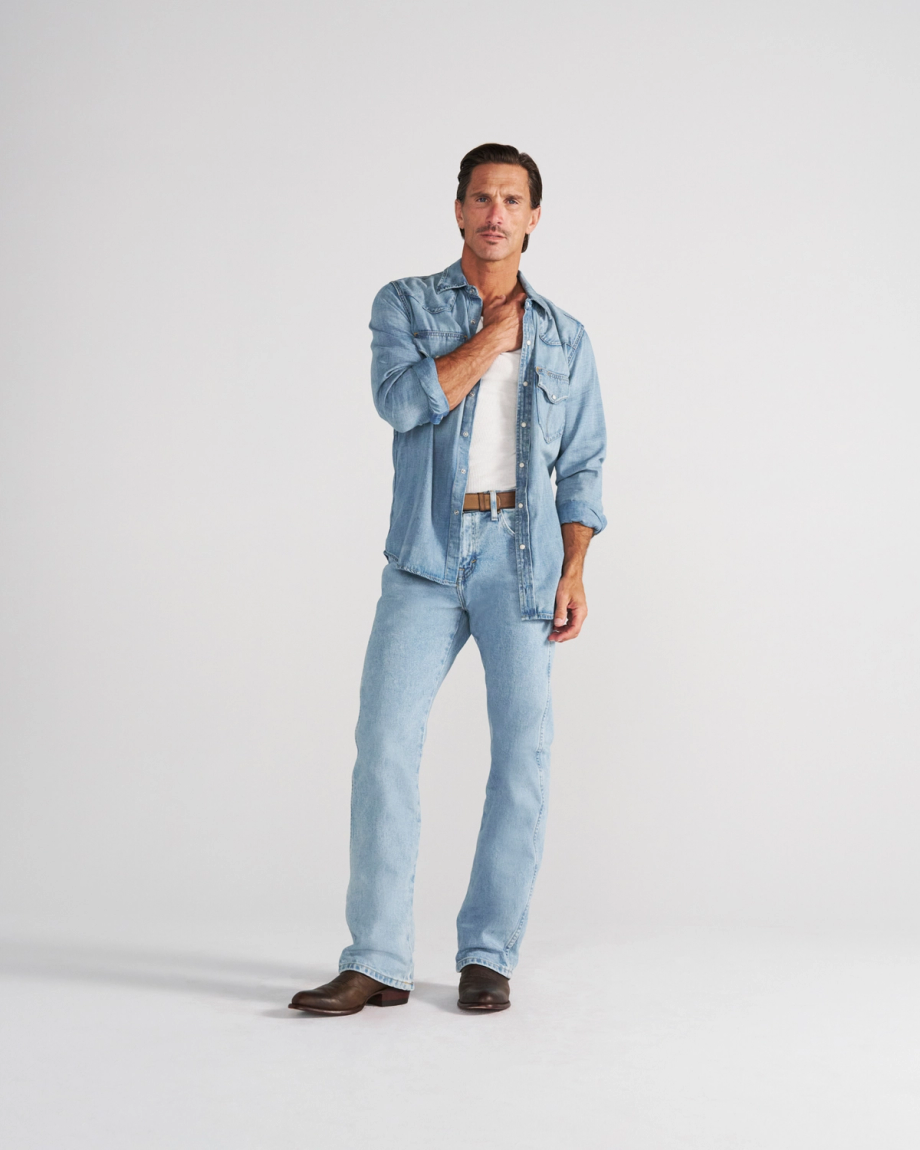 A man wearing a light-wash denim shirt over a white tank top, light-wash blue jeans, and brown leather boots.
