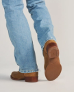 The Buck in Copper Cowhide. A person wearing light blue jeans and brown shoes—crafted from Buck Cowhide—is walking away; the sole of one shoe, showing a tread pattern and branding, is visible.