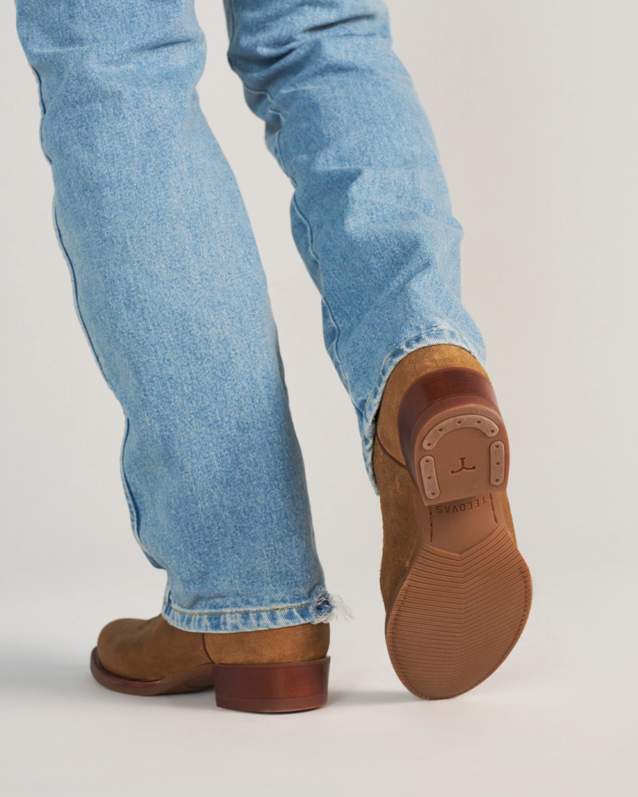 The Buck in Copper Cowhide. A person wearing light blue jeans and brown shoes—crafted from Buck Cowhide—is walking away; the sole of one shoe, showing a tread pattern and branding, is visible.