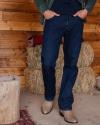 A person stands indoors wearing a green shirt, dark blue jeans, and brown cowboy boots, with hands in pockets. Hay bales, a wooden wall, and a tree stump are in the background.