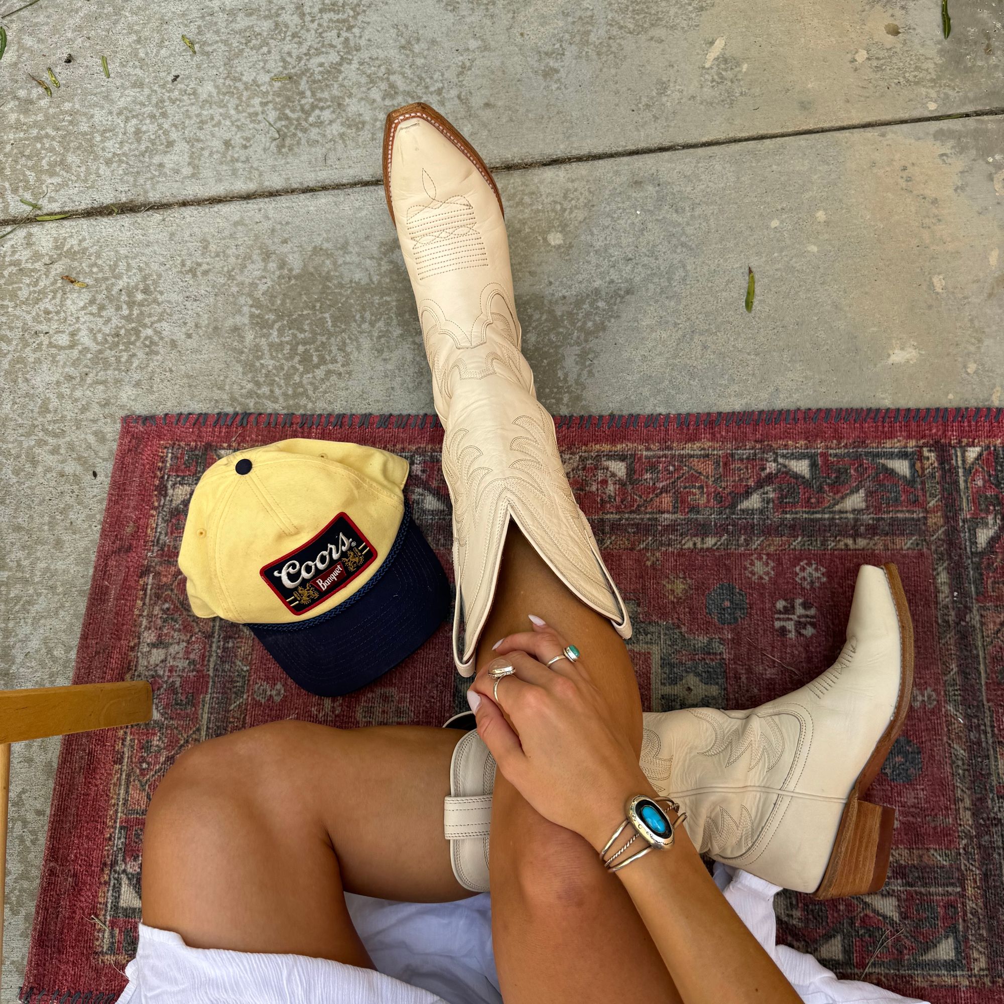Person wearing cream cowboy boots and turquoise jewelry sits on a red patterned rug next to a yellow and navy "Coors" hat. Only their legs and hands are visible.