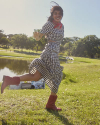 Woman in a checked dress and red boots joyfully runs through a grassy park near a pond on a sunny day.