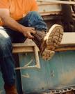 LongHaul Waterproof Square Composite Toe Work Boot in Saddle/Fossil Cowhide. A person wearing jeans and an orange shirt sits on metal steps, pulling on a LongHaul Waterproof Square Composite Toe Work Boot Cowhide.