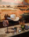 A white cowboy hat, leather belts, a green glass goblet with feathers, and other decorative items displayed on a counter with a mural in the background.