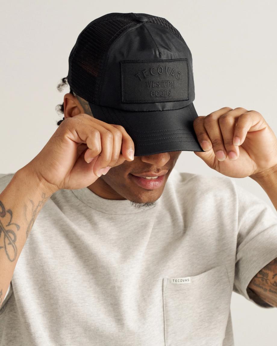 A person wearing a light gray t-shirt and a black Tecovas cap adjusts the brim of the hat with both hands.