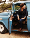 Man in denim sitting in the open door of a vintage blue truck, looking away thoughtfully, with string lights in the background.