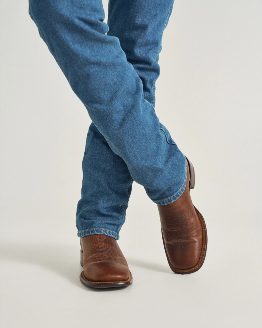 The Doc in Burnt Umber / Blue Cowhide. Person wearing blue jeans and The Doc Cowhide brown leather cowboy boots stands with legs crossed against a plain background.