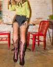The Annie in Berry Cowhide. Person wearing a green western shirt, black fringed shorts, and purple cowboy boots sits on a red chair next to a table featuring The Annie Cowhide in a rustic setting.
