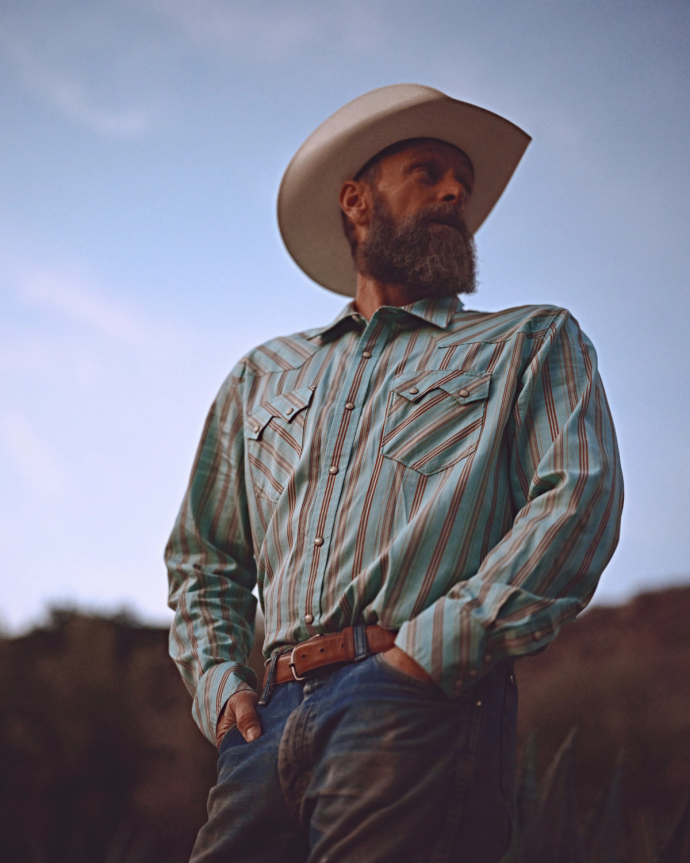 A bearded man in a cowboy hat and striped shirt stands outdoors with hands in his pockets, looking to the side.