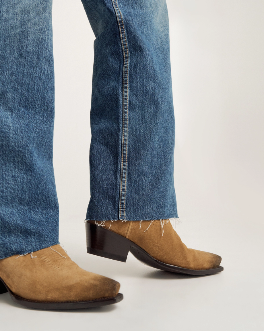 Close-up of a person wearing blue jeans with frayed hems and brown suede cowboy boots on a neutral background.