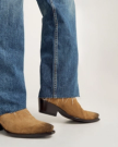 Close-up of a person wearing blue jeans with frayed hems and brown suede cowboy boots on a neutral background.