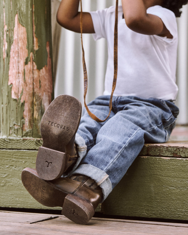 Child in jeans and a white shirt sits on a wooden step with legs extended, wearing brown Tecovas boots and holding a camera strap.