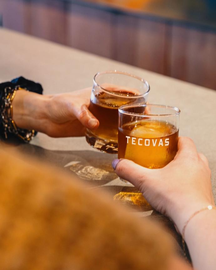 Two people clink glasses of amber-colored drinks. One glass displays the word "Tecovas." The glasses appear to be resting on a bar or tabletop.