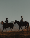 Two people in cowboy hats ride horses in silhouette against a mountain landscape at sunset.