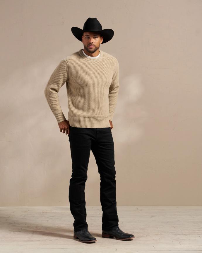 A man wearing a beige sweater, black trousers, black boots, and a black cowboy hat stands against a plain background.