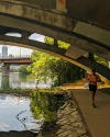A shirtless person jogs along a pathway under a concrete bridge by a river, with trees and city buildings visible in the background.