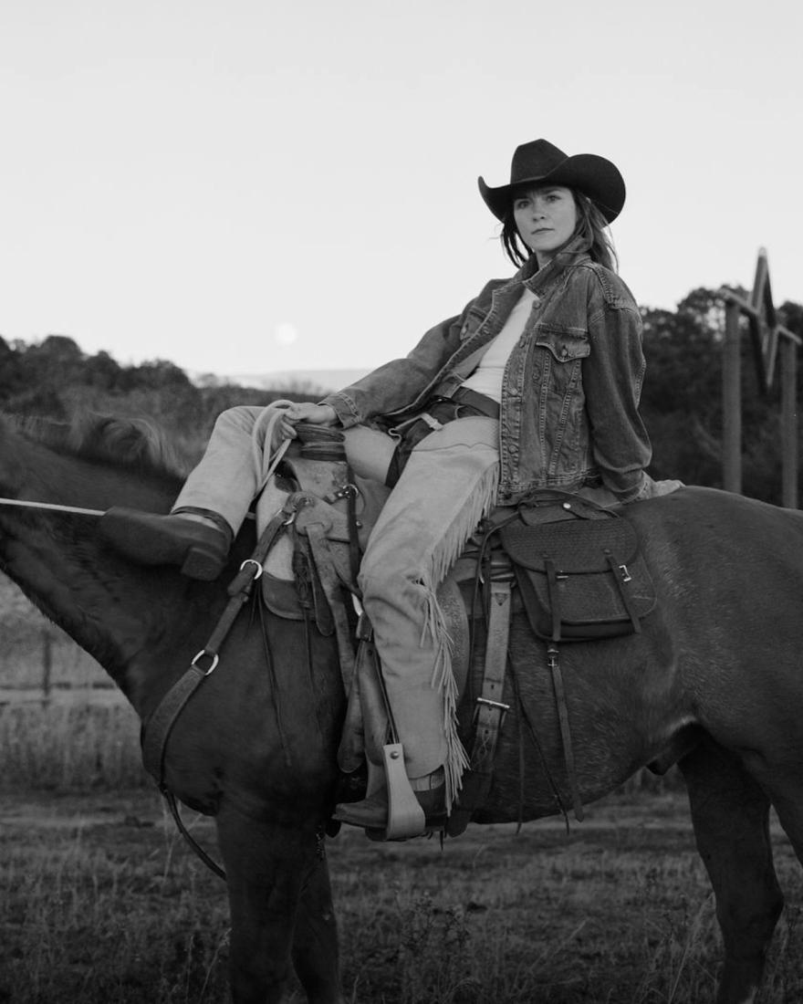 A person wearing a cowboy hat and denim clothing sits on a saddled horse outdoors, with trees and fields in the background.