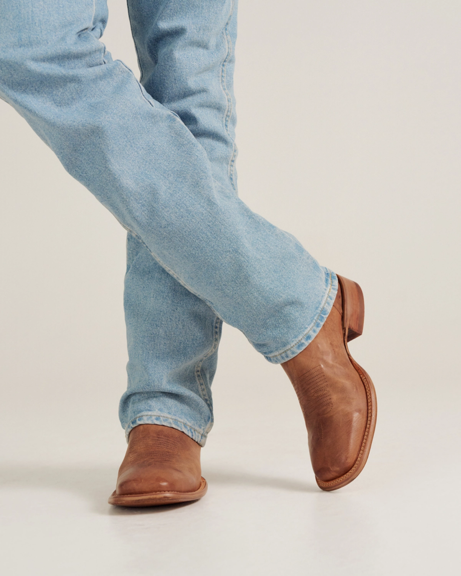 The Doc in Scotch Goat. Person wearing light blue jeans and brown leather boots from The Doc Goat stands with legs crossed on a plain background.