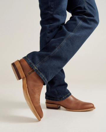 A person wearing blue jeans and brown leather boots with stacked heels stands with one leg crossed over the other against a plain background.