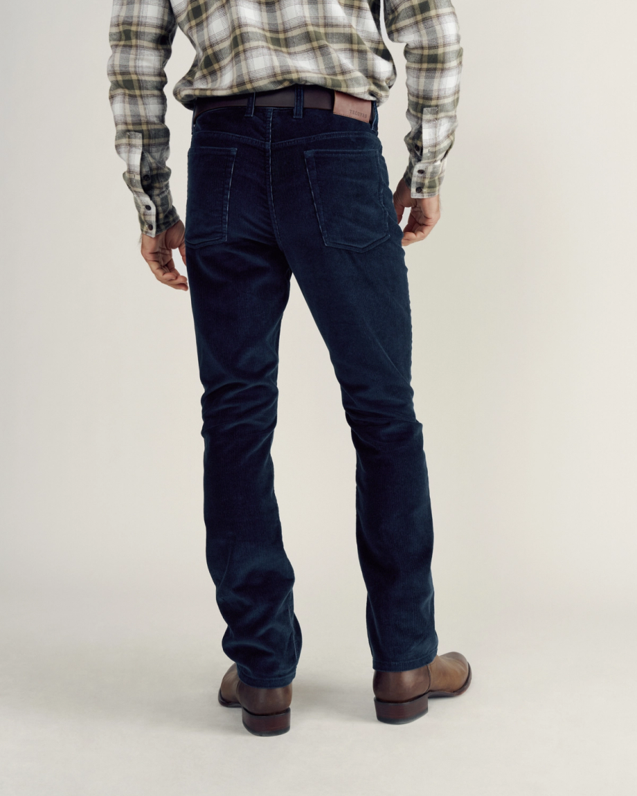 A person stands facing away, wearing a plaid shirt, dark jeans, a brown belt, and brown boots against a plain background.