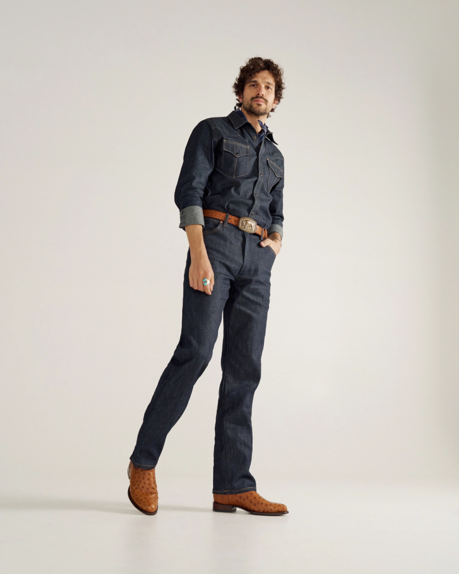 A man stands against a plain background wearing a dark denim shirt and jeans, a brown belt, brown cowboy boots, and a turquoise ring.