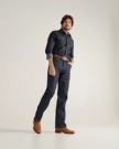 A man stands against a plain background wearing a dark denim shirt and jeans, a brown belt, brown cowboy boots, and a turquoise ring.