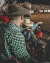 A cowboy in a striped shirt prepares for a rodeo event, adjusting his grip on a bull rope. Several people in cowboy hats surround him in a bustling arena.