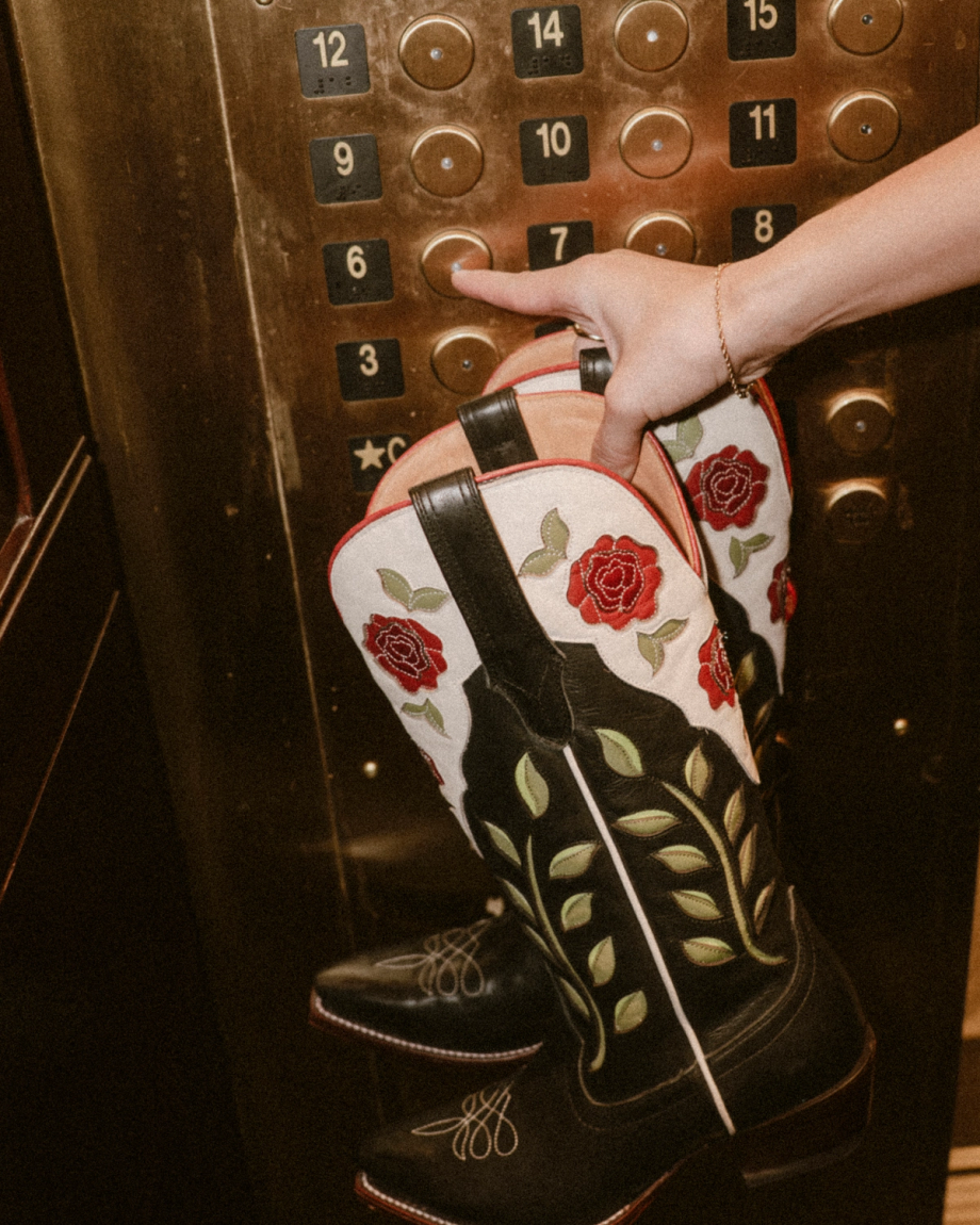A hand holds The Annie Cowhide black and white cowboy boots with striking red rose embroidery—Midnight Roses—in an elevator, pressing a floor button.
